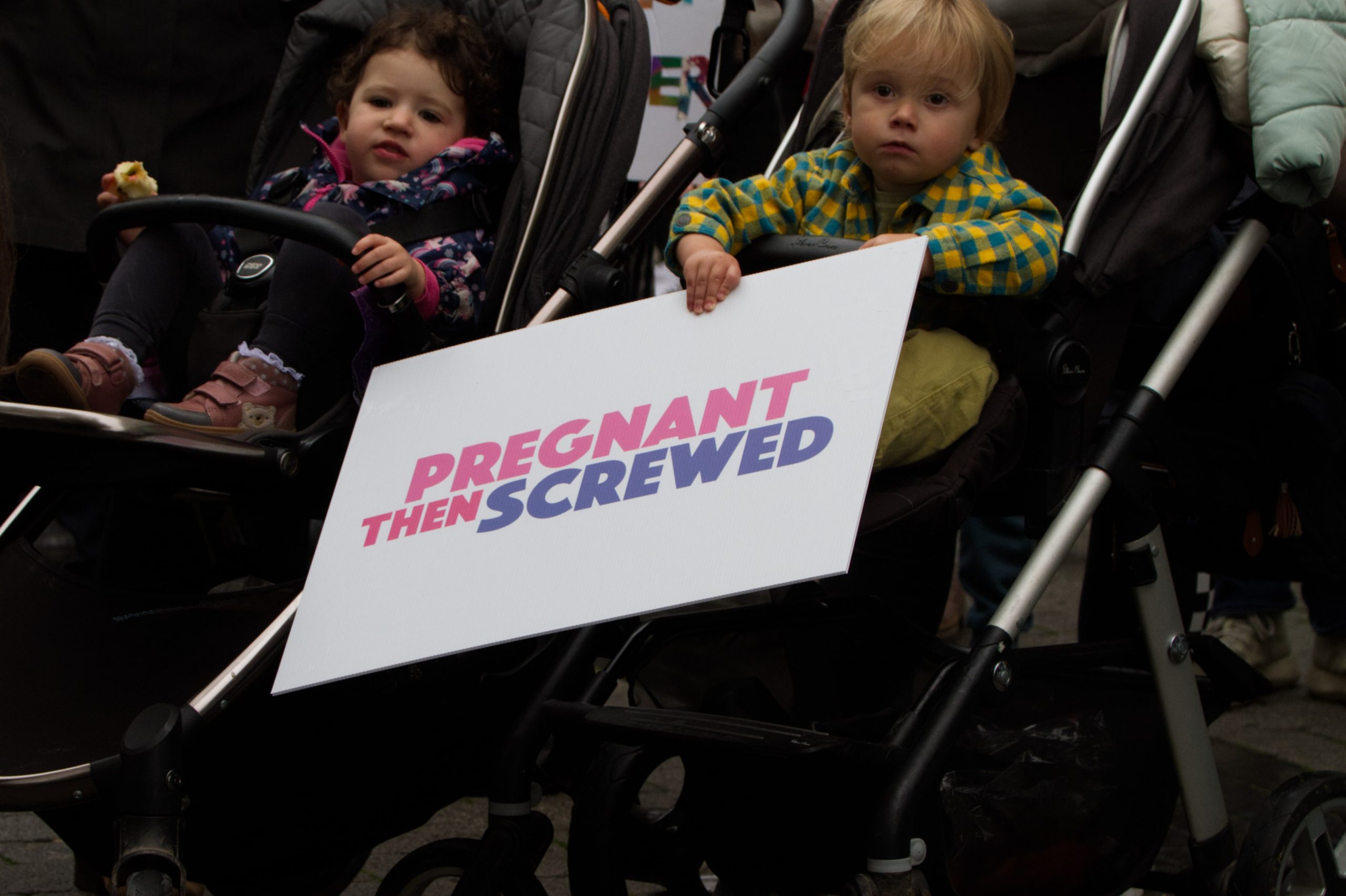 Alt text: child in buggy holding a Pregnant Then Screwed placard