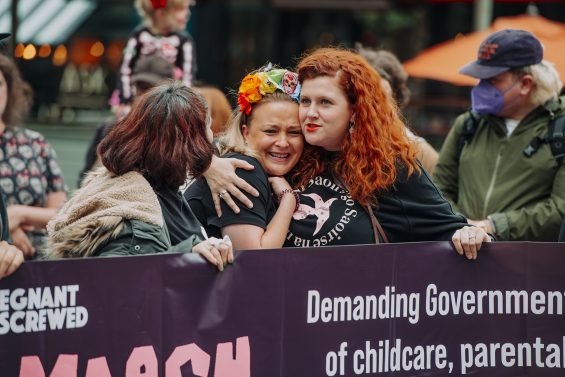 Alt text: women hugging behind a Pregnant Then Screwed protest banner at March of the Mummies Cardiff