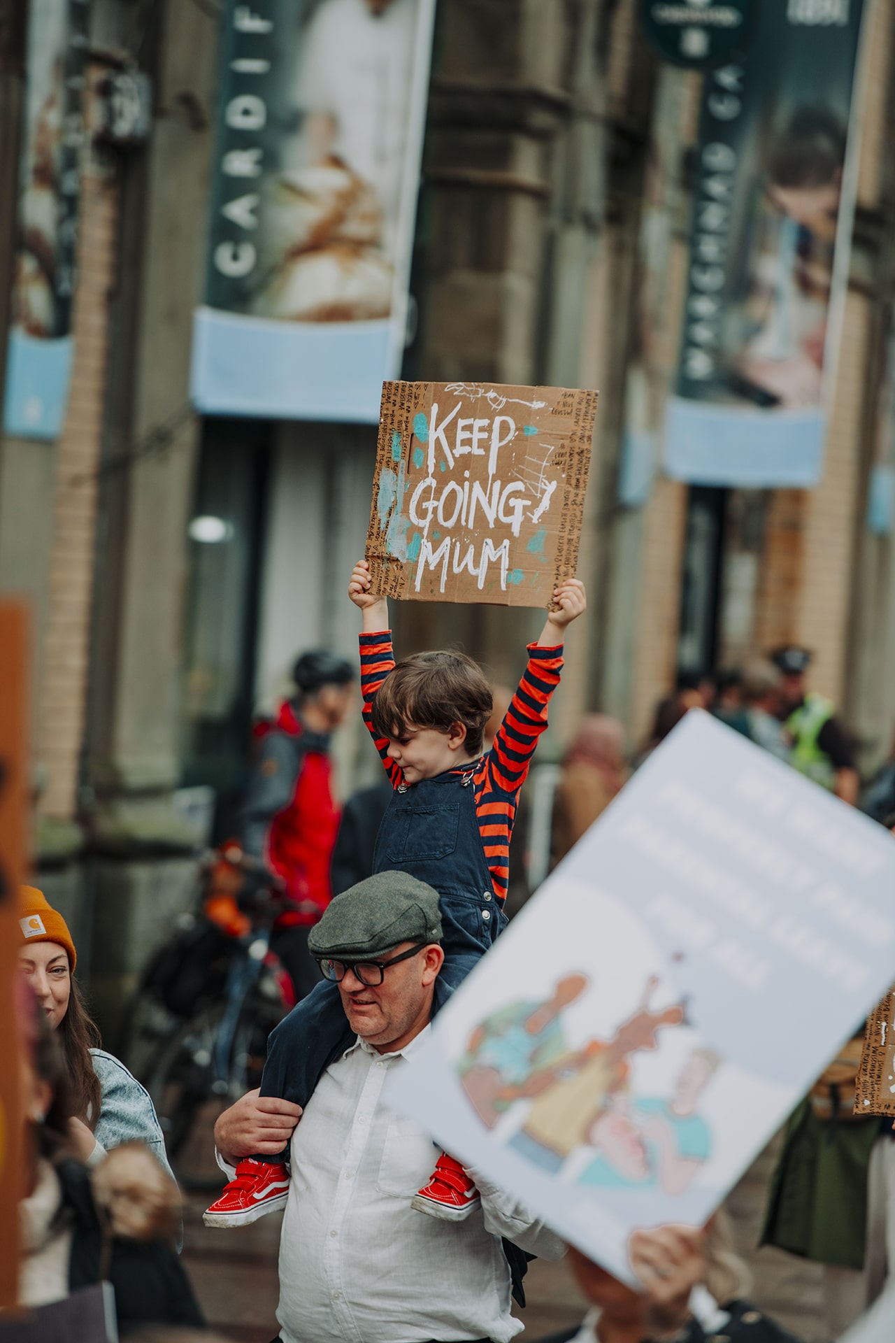 Alt text: xhild on his fathers' shoulders holding up a sign saying 