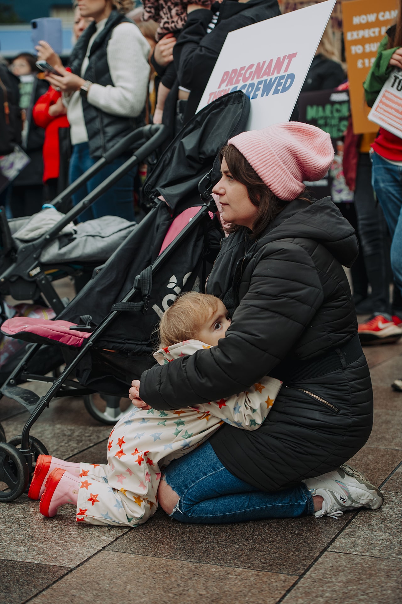 Alt text: woman crouched on the ground at a Match of the Mummies protest hugging her child