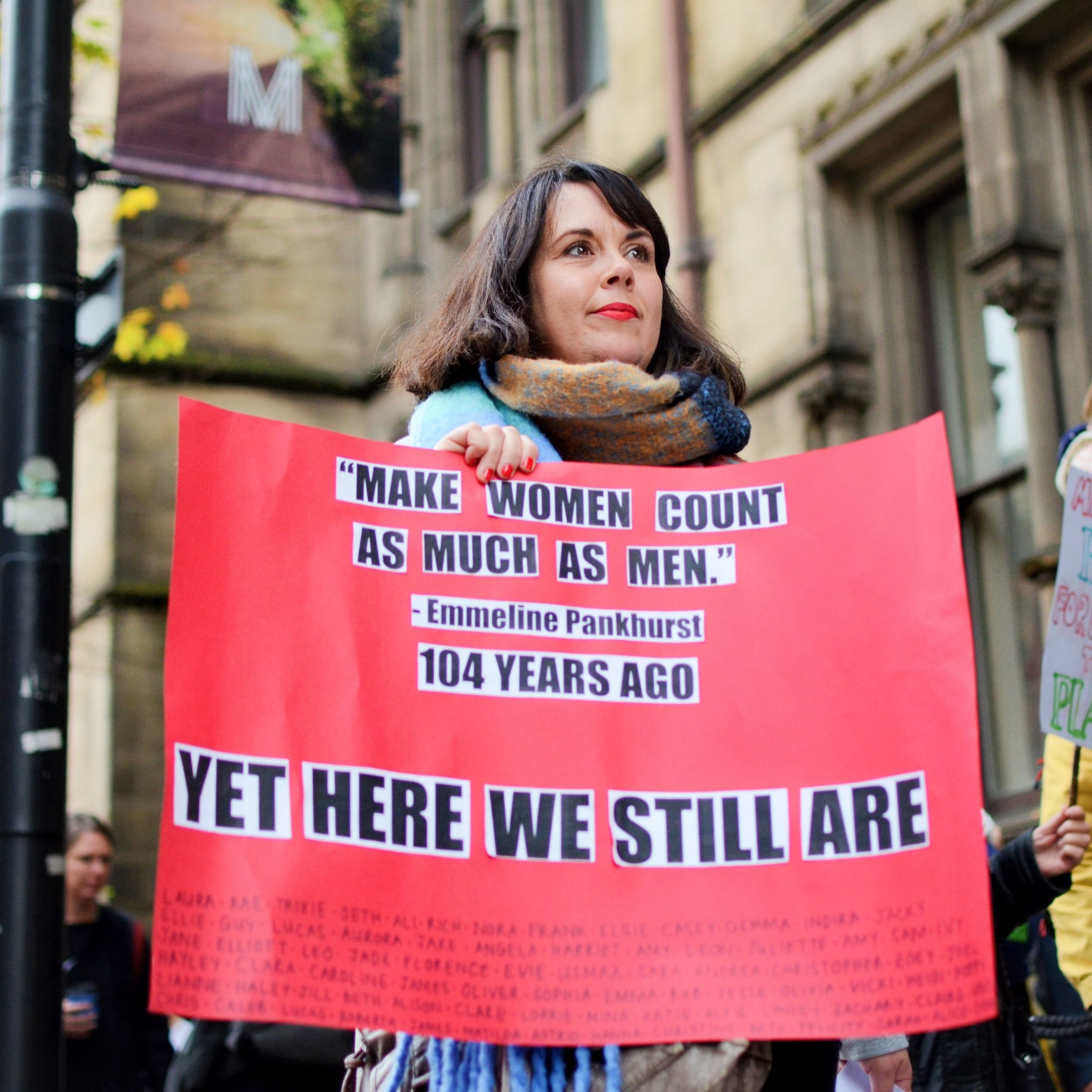 Woman holding a red sign that reads 'Make women count as much as men' - yet here we still are