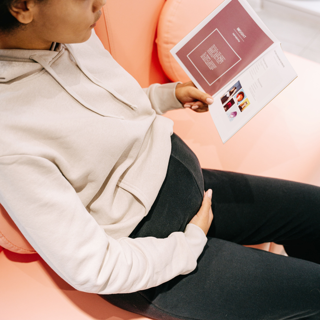 Alt text: Pregnant brown woman, istting on the sofa with her hadn pn her belly reading a pamphlet