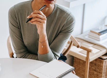 Alt text: women in grey jumper sitting bu a notebook with her pen pressed to her chin in thought